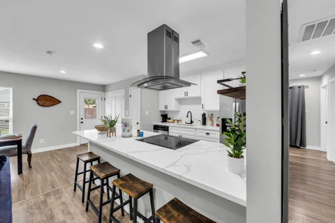 Bright white kitchen with an island, black stools, a stainless range hood, and adjoining dining space